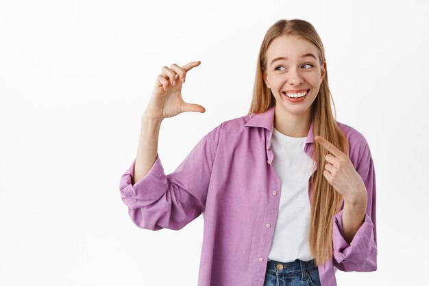 Mujer rubia sosteniendo algo pequeño y finito Chica riendo mientras señala su mano mostrando una cosa diminuta de tamaño pequeño sonriendo de pie contra el fondo blanco