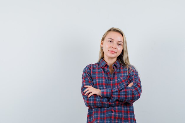 Mujer rubia sosteniendo los brazos cruzados y sonriendo con camisa a cuadros y luciendo atractiva, vista frontal.