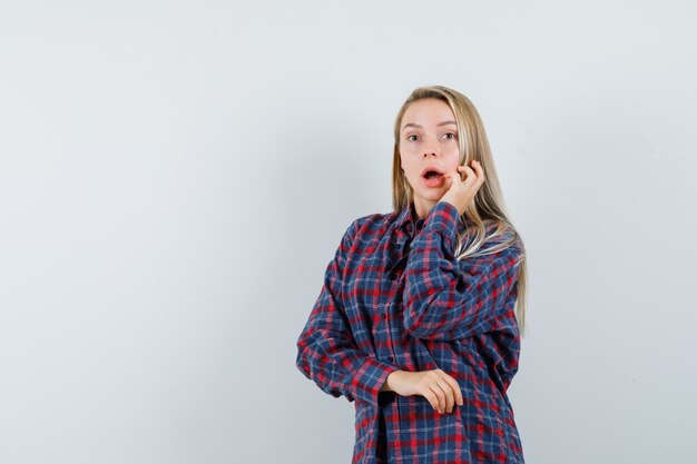 Mujer rubia sosteniendo una mano cerca de la boca y posando a la cámara con camisa a cuadros y mirando sorprendido. vista frontal.