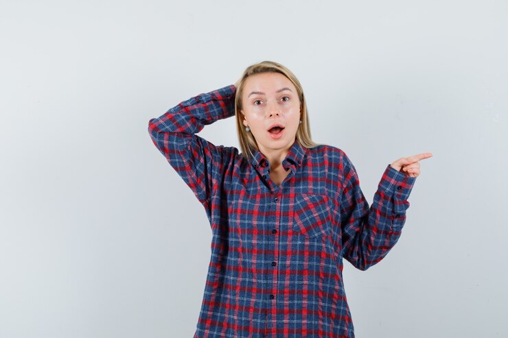 Foto gratuito mujer rubia sosteniendo la mano detrás de la cabeza y apuntando a la derecha con el dedo índice en camisa a cuadros y mirando sorprendido, vista frontal.