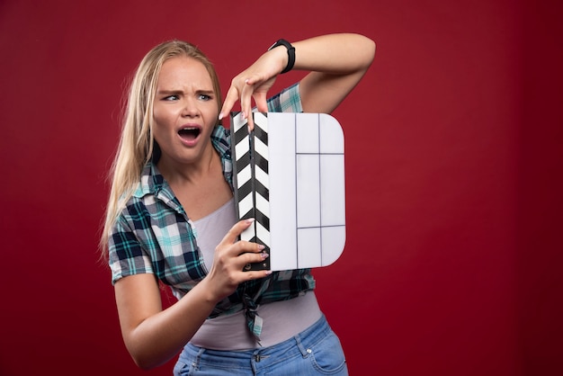 Foto gratuita mujer rubia sosteniendo una placa de azote de producción de películas y parece confundida y cansada.