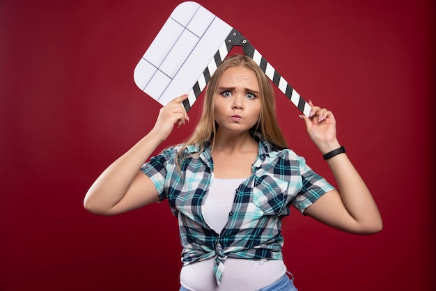 Foto gratuita mujer rubia sosteniendo una placa de azote de producción de películas y parece confundida y cansada.