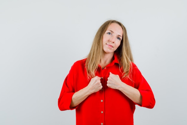 Foto gratuita mujer rubia sosteniendo los puños en el pecho con camisa roja y mirando confiada.