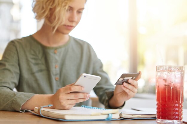 Mujer rubia sosteniendo el teléfono y la tarjeta de crédito en la cafetería mientras toma una copa