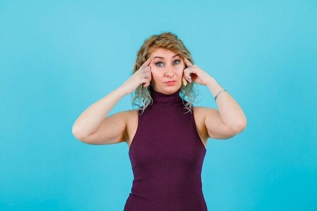 Mujer rubia sostiene los dedos índices en las sienes sobre fondo azul.