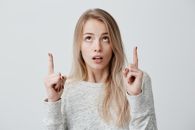 Mujer rubia en suéter con expresión de la cara asombrada manteniendo los dedos índices apuntando hacia arriba. Linda mujer caucásica mostrando algo por encima de la cabeza, haciendo gesto con el dedo índice. Choque y asombro