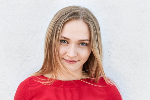 Mujer rubia en suéter rojo mirando con confianza a la cámara con mirada misteriosa.