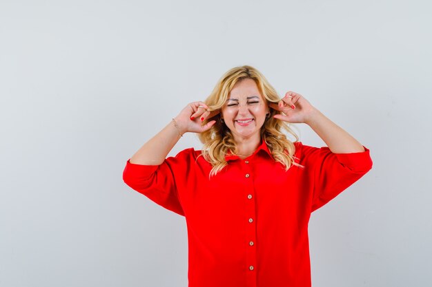 Mujer rubia tapando los oídos con los dedos índices en blusa roja y mirando acosado