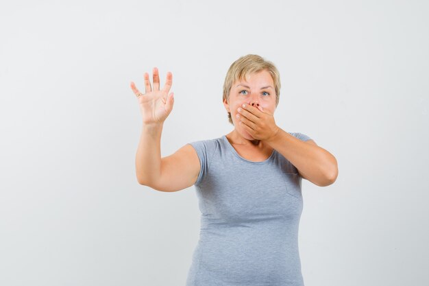 Mujer rubia tapándose la boca con la mano en una camiseta azul claro y mirando sorprendido, vista frontal.