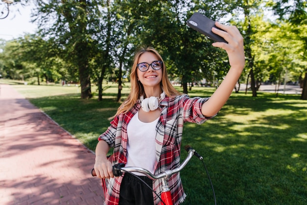 Mujer rubia tímida en gafas con teléfono para selfie en buen día de verano. Chica bastante caucásica posando con bicicleta roja.
