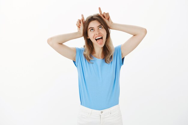 Mujer rubia tonta despreocupada mostrando lengua y cuernos por encima de la cabeza