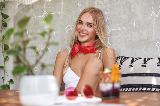 Mujer rubia con top blanco y pañuelo rojo posando en un café