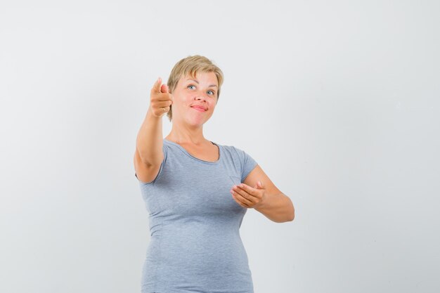 Mujer rubia tratando de mostrar algo en una camiseta azul claro y luciendo optimista, vista frontal.