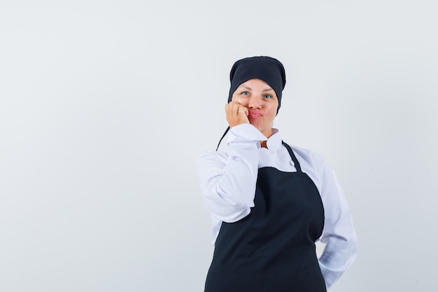 Mujer rubia en uniforme de cocinero negro apoyando la mejilla en la mano, pensando en algo y luciendo bonita