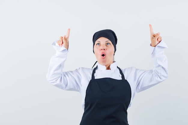 Foto gratuita mujer rubia en uniforme de cocinero negro apuntando hacia arriba con los dedos índices y luciendo bonita