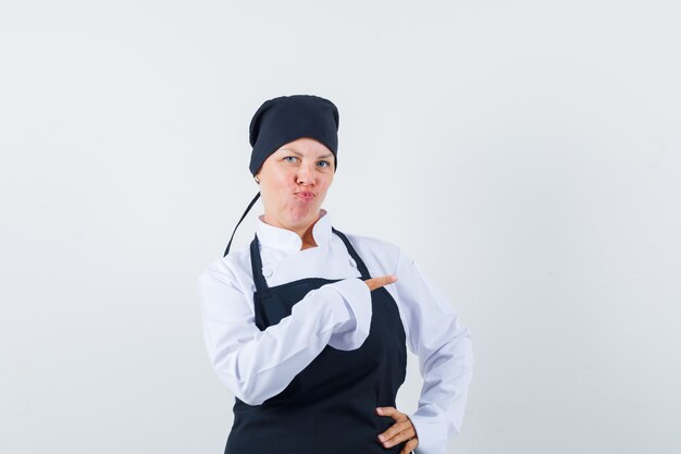 Mujer rubia en uniforme de cocinero negro apuntando a la derecha con el dedo índice, labios curvos y bonita