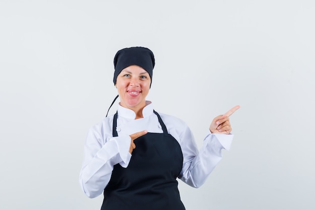 Mujer rubia en uniforme de cocinero negro apuntando a la derecha con los dedos índices y luciendo bonita