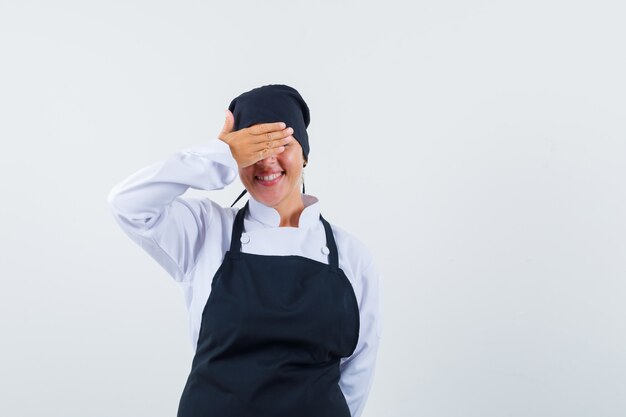 Mujer rubia en uniforme de cocinero negro cubriendo los ojos con las manos y mirando tímido