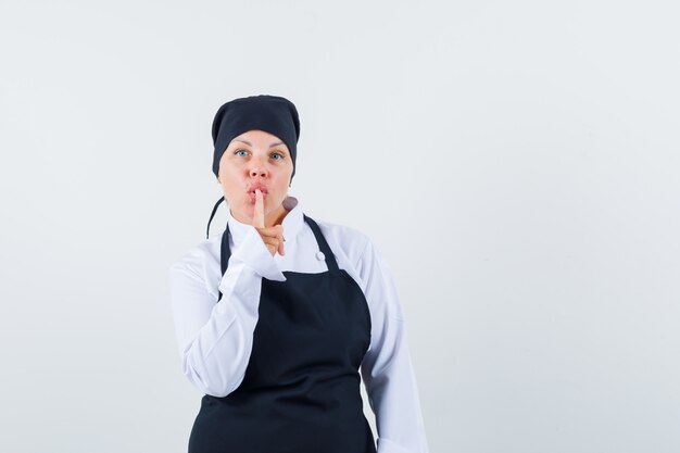 Mujer rubia en uniforme de cocinero negro mostrando gesto de silencio y luciendo bonita