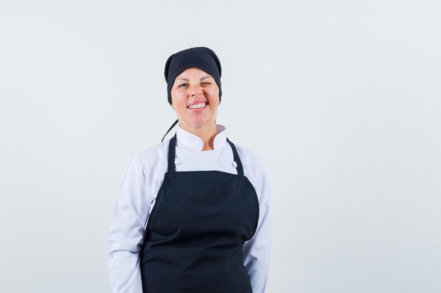 Mujer rubia en uniforme de cocinero negro de pie derecho y posando al frente y mirando bastante