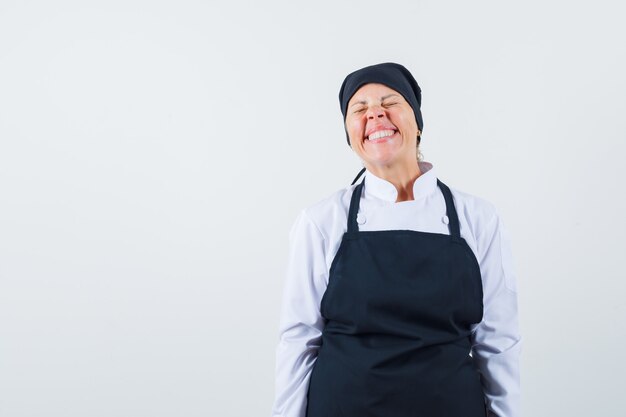 Mujer rubia en uniforme de cocinero negro de pie, sonriendo con gracia y posando a la cámara y mirando bonita, vista frontal.