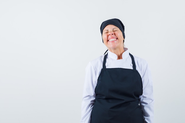 Mujer rubia en uniforme de cocinero negro de pie, sonriendo con gracia y posando a la cámara y mirando bonita, vista frontal.