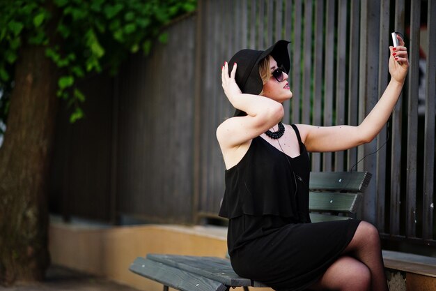 Mujer rubia con vestido negro gafas de sol collares y sombrero sentado en un banco con teléfono móvil a mano y auriculares