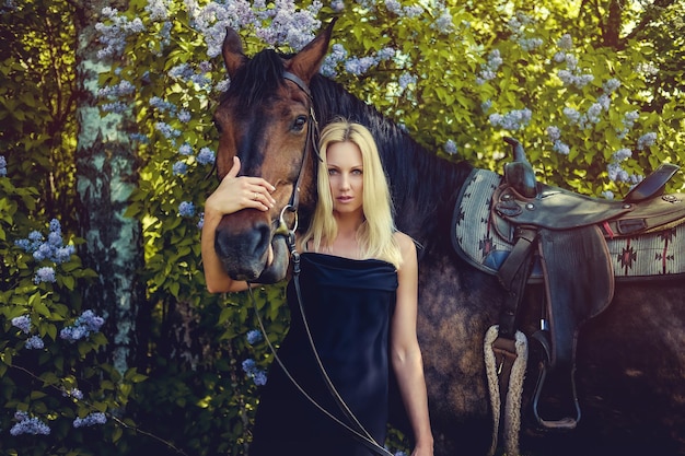 Mujer rubia en vestido de noche negro posando con caballo marrón.