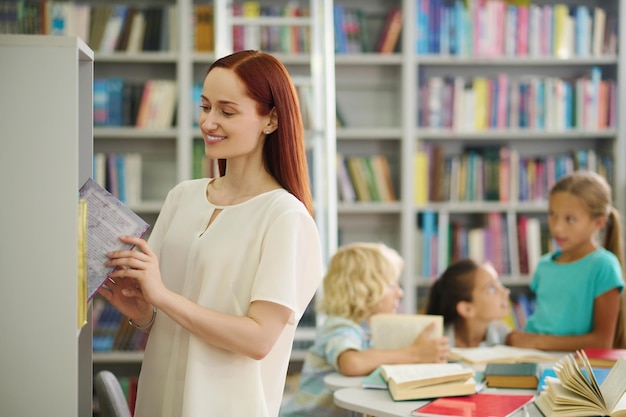 Mujer sacando un libro cerca de una estantería y niños detrás