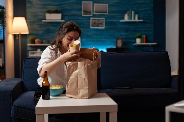 Mujer en la sala de estar preparándose para la cena en el sofá desempacando papas fritas y una sabrosa hamburguesa de una bolsa de papel para llevar. Persona que come comida chatarra de hamburguesas y cerveza embotellada sentada en el sofá.