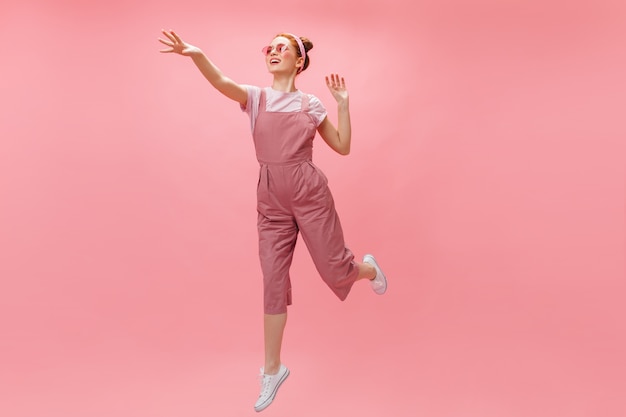 mujer saltando sobre fondo rosa. Tiro de cuerpo entero de mujer pelirroja en traje brillante y gafas.