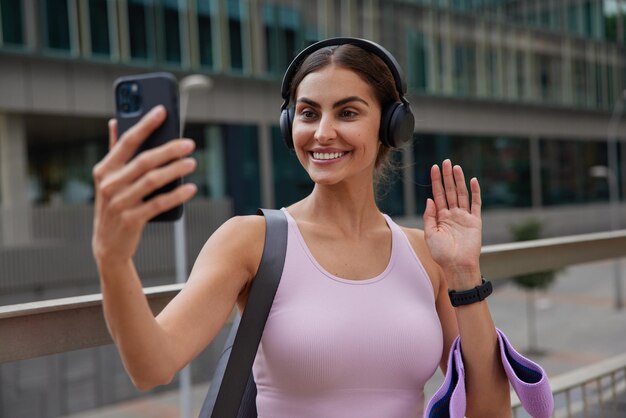 mujer saluda con la palma hace una videollamada a través de un teléfono inteligente lleva una banda de resistencia y karemat usa auriculares inalámbricos posa al aire libre para entrenar al aire libre da un tutorial