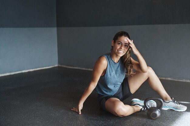 Mujer satisfecha jadeando sesión de entrenamiento de entrenamiento productivo Atractiva deportista limpiar el sudor de la frente sonriendo complacido descansar sentarse gimnasio piso terminar entrenamiento sesión de entrenamiento tren pesas rusas