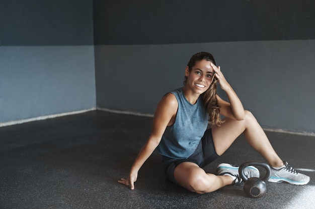 Mujer satisfecha jadeando sesión de entrenamiento de entrenamiento productivo Atractiva deportista limpiar el sudor de la frente sonriendo complacido descansar sentarse gimnasio piso terminar entrenamiento sesión de entrenamiento tren pesas rusas