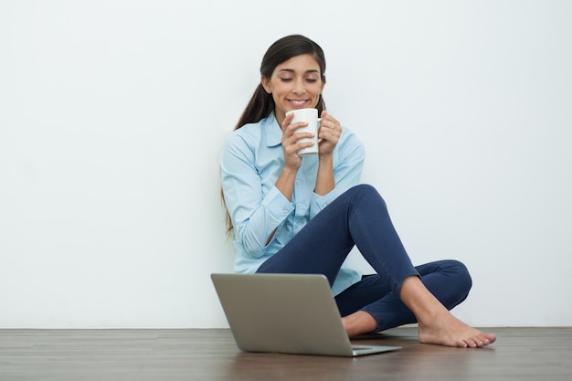Mujer satisfecha que bebe té en suelo con el ordenador portátil