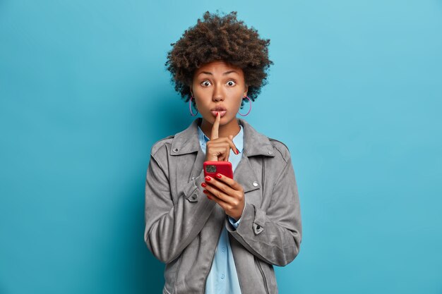 Mujer secreta sorprendida con cabello rizado, piel oscura, hace gesto de silencio, sostiene el teléfono móvil, crea un nuevo perfil en las redes sociales, cuenta información secreta o confidencial, posa sobre una pared azul