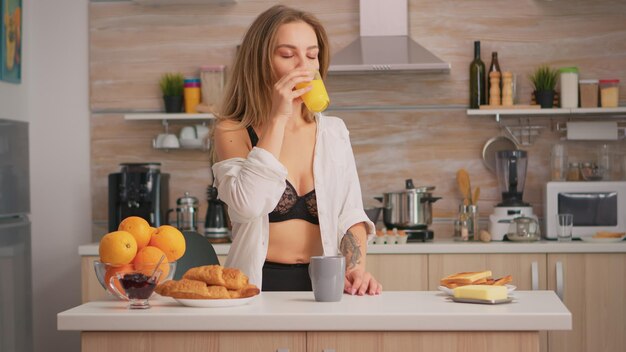 Mujer seductora en ropa interior sexy disfrutando de la mañana bebiendo un vaso de jugo de naranja fresco sentado en la cocina. Jovencita blode con tatuajes vistiendo lencería negra refrescante domingo por la mañana