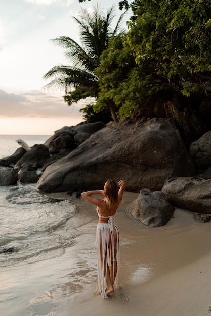 Mujer seductora con vestido de ganchillo boho blanco posando en la playa. Foto de moda de verano y vacaciones tropicales. De longitud completa.