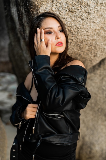 Mujer segura con labios rojos en chaqueta de cuero negro de moda y vestido negro sensual posando sobre fondo rocoso