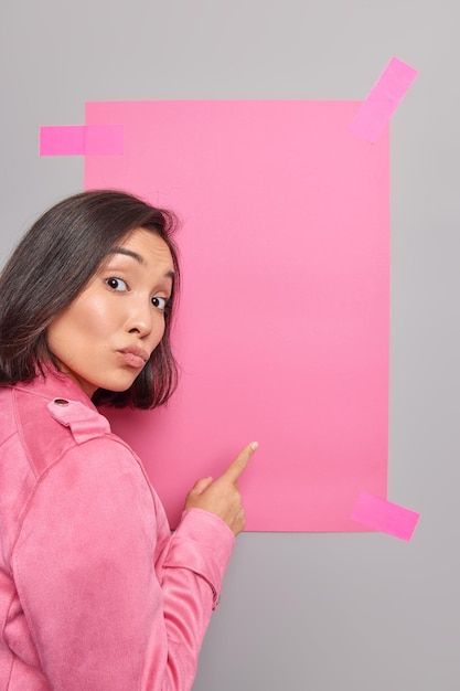 Foto gratuita mujer señala con el dedo índice a la pancarta de papel rosa vacía sugiere colocar viste chaqueta rosa atrae su atención hacia algo.