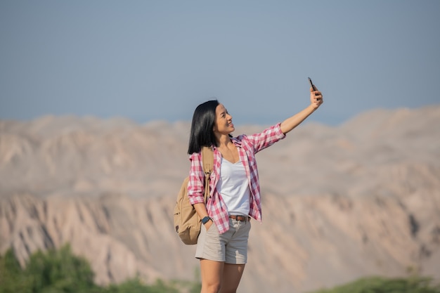 Mujer de senderismo en las montañas de pie en la cresta rocosa de la cumbre con mochila y poste mirando el paisaje, mujer feliz haciendo autorretrato en las montañas