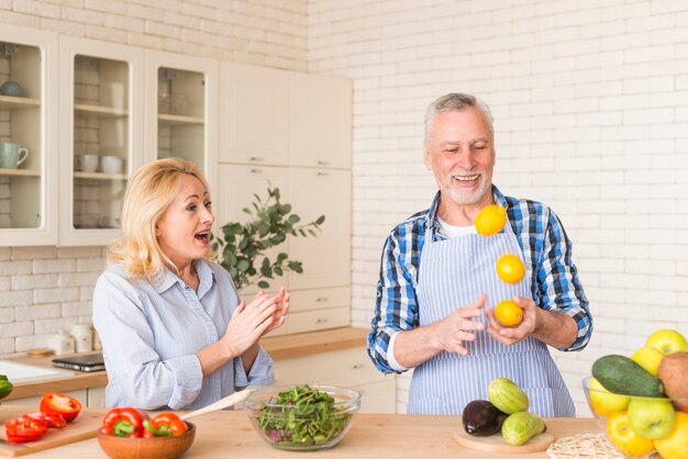 Mujer Senior aplaudiendo mientras su esposo hace malabares con naranjas enteras en la cocina