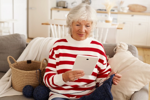 Mujer senior atractiva en sudadera blanca roja relajante en el interior, sentado en el sofá con hilo y agujas, tejido, tableta digital usng para compras en línea. Personas mayores, jubilación, tecnología moderna