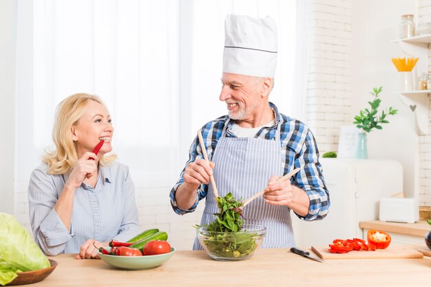 Mujer Senior mordiendo el chile rojo mirando a preparar la ensalada en la cocina