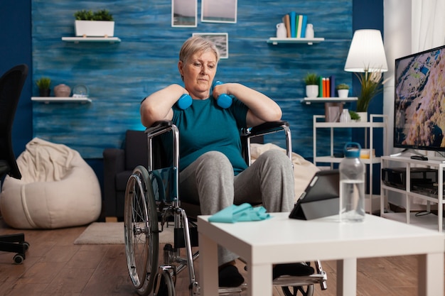 Mujer senior no válida en silla de ruedas viendo ejercicio corporal de gimnasio en tableta en la sala de estar
