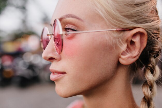 mujer sensual bronceada mirando a distancia con expresión de la cara interesada. Retrato al aire libre de mujer rubia con trenzas con gafas de sol rosas redondas.