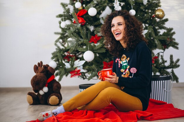 Mujer sentada por el árbol de navidad y bebiendo té