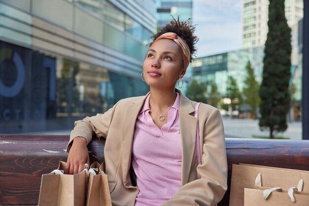 Mujer sentada en un banco con bolsas de la compra alrededor vestida con ropa de moda centrada arriba plantea contra el entorno urbano piensa en algo descansa después de caminar