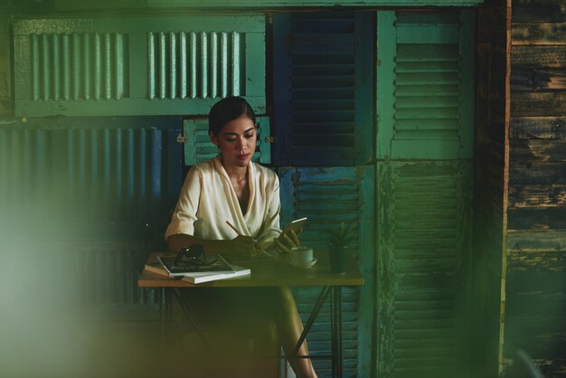 Mujer sentada en la cafetería, mirando el teléfono inteligente y escribiendo en el cuaderno