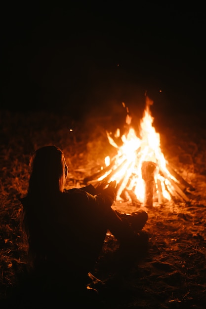 Mujer sentada y calentándose cerca de la hoguera en el bosque nocturno